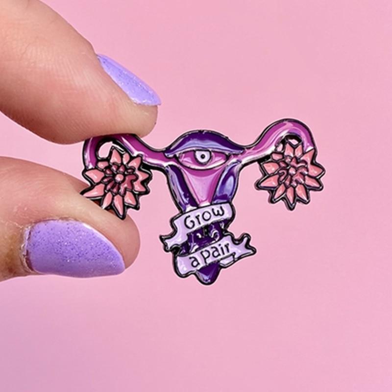 Hand holding a purple uterus enamel pin with pink flowers and “GROW A PAIR” banner