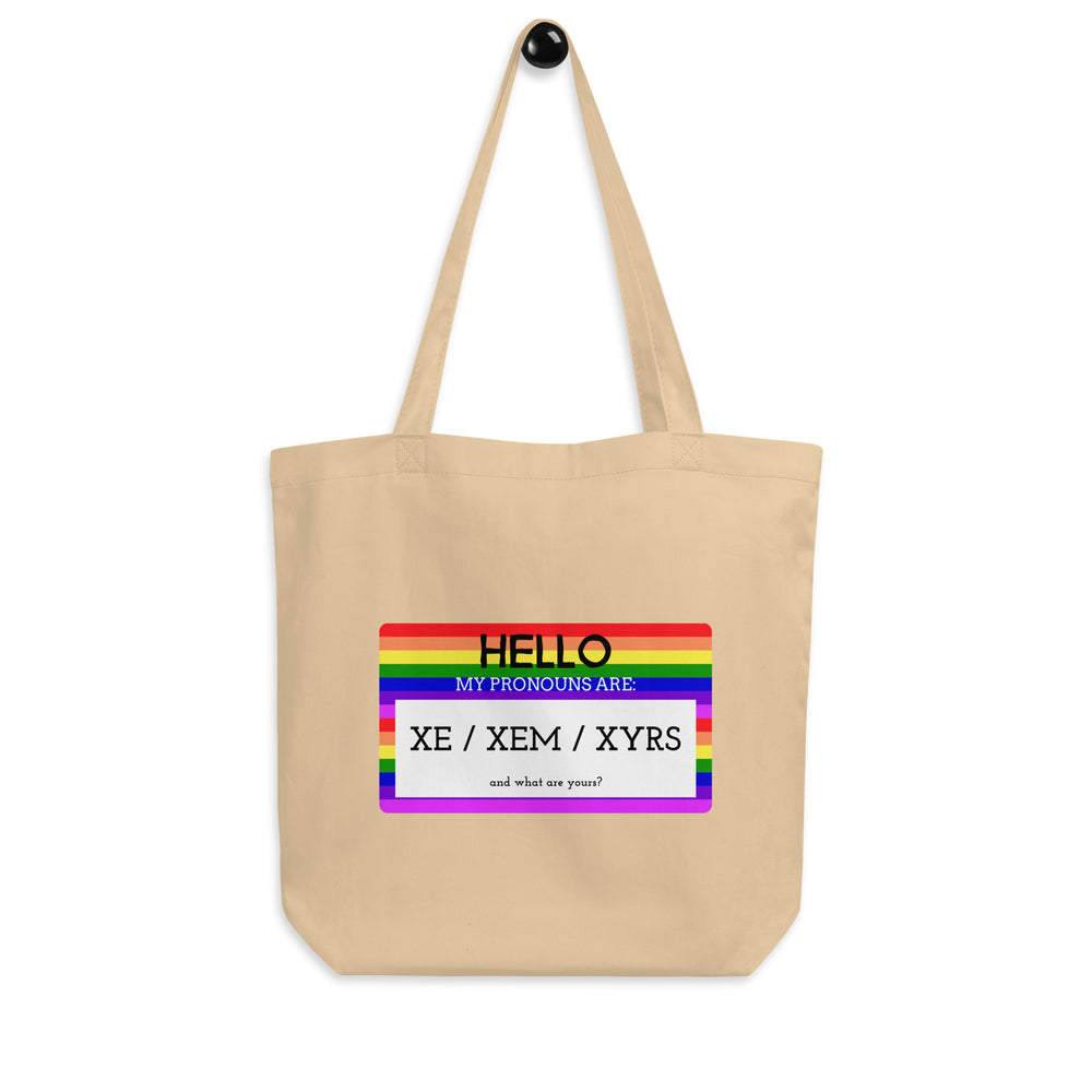 Front view of a beige tote bag with a rainbow label reading “Hello, my pronouns are: xe / xem / xyrs.”