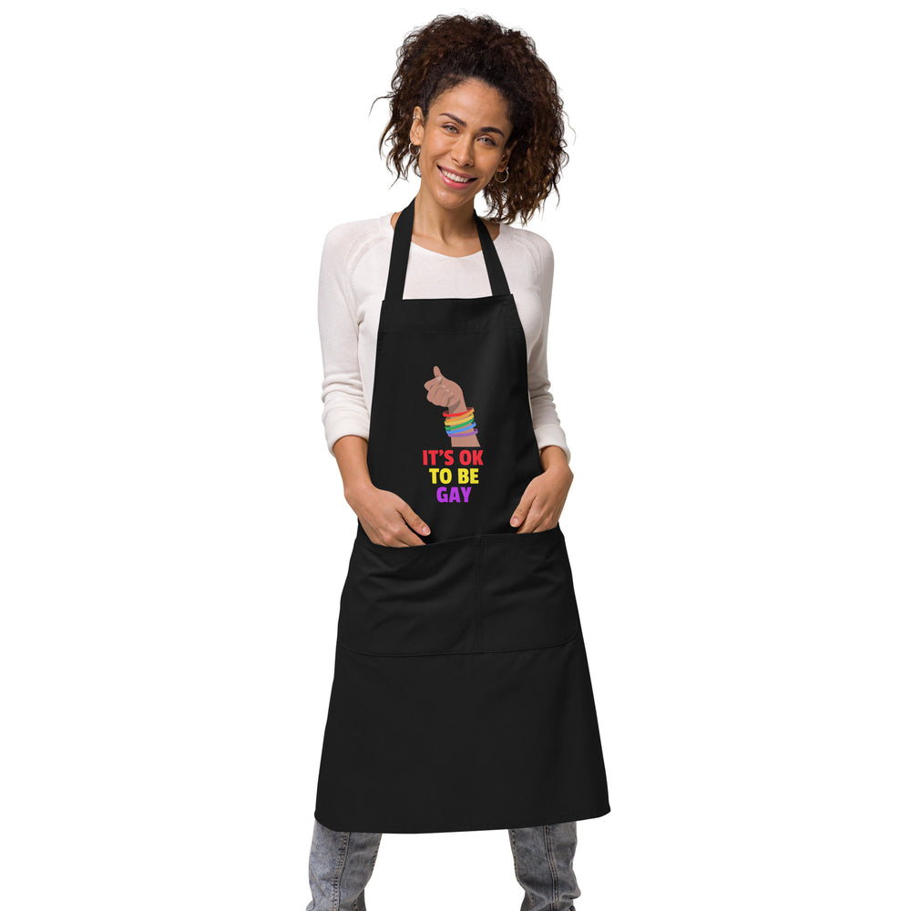 Front view of a model wearing a black apron reading “IT’S OK TO BE GAY” with a rainbow-cuffed fist
