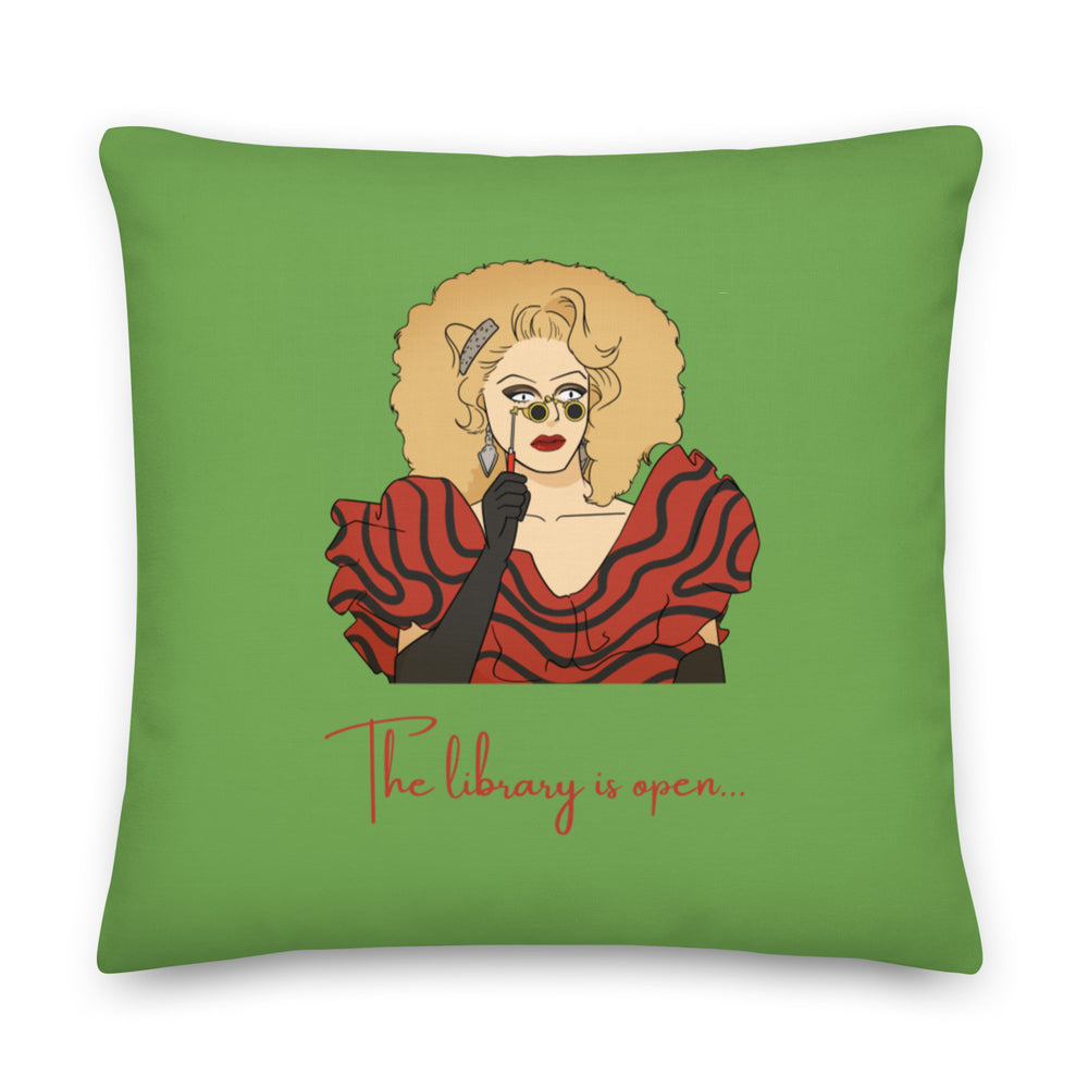 Green square pillow with a drag performer illustration and red text reading “The library is open...”