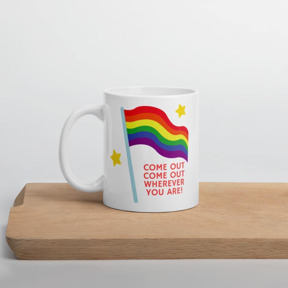 White mug on a wooden stand with a rainbow flag and “COME OUT COME OUT WHEREVER YOU ARE!”