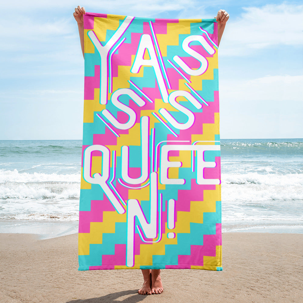 Colorful beach towel reading “Yasss Queen!” held upright on a sandy beach