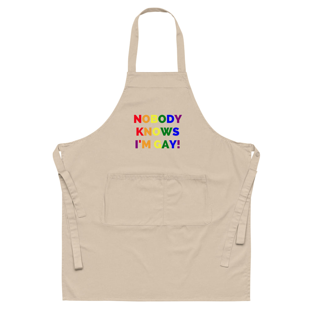Front view of a beige apron reading “NOBODY KNOWS I'M GAY!” in rainbow letters
