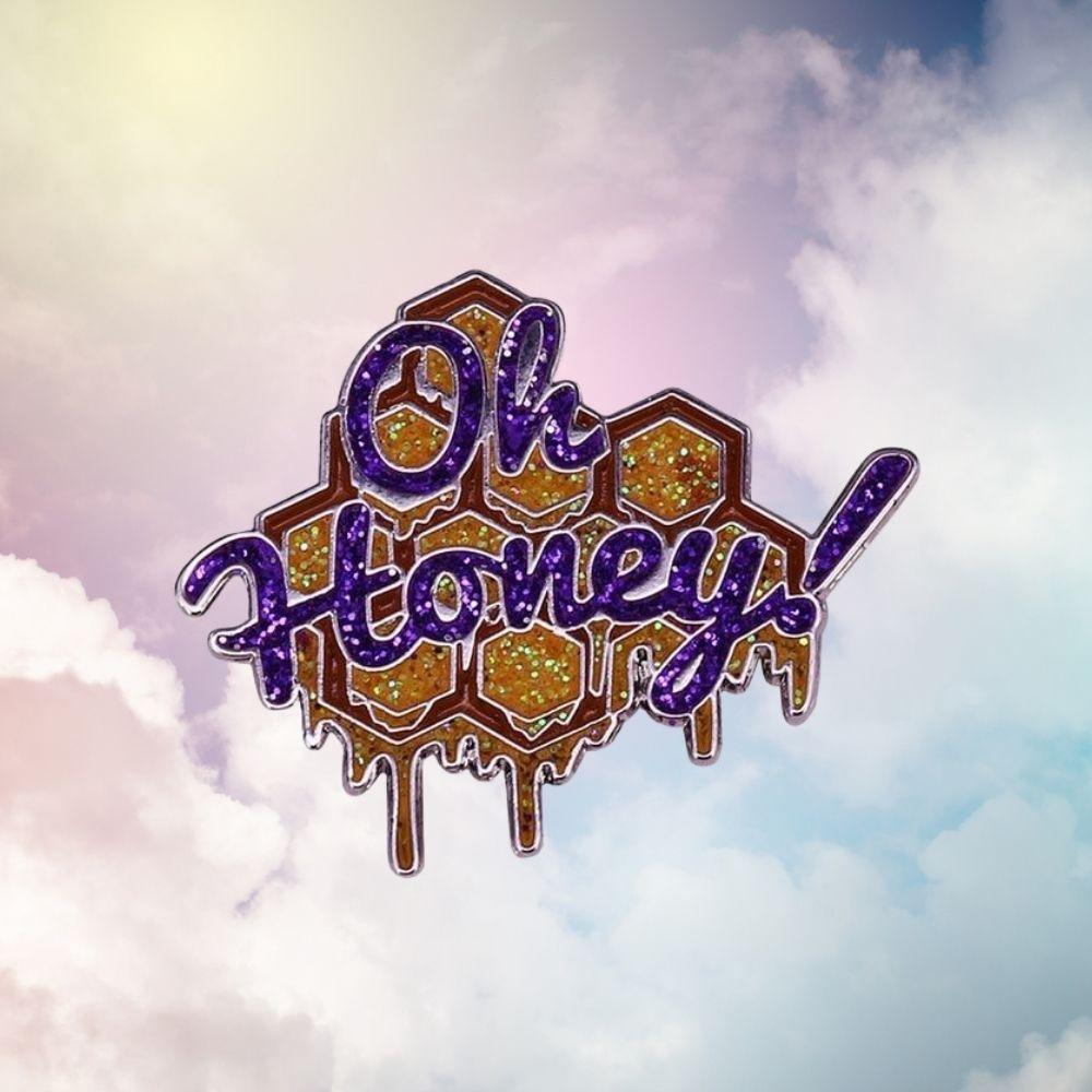 Close-up of a glittery purple and gold enamel pin reading “Oh Honey” over honeycomb shapes