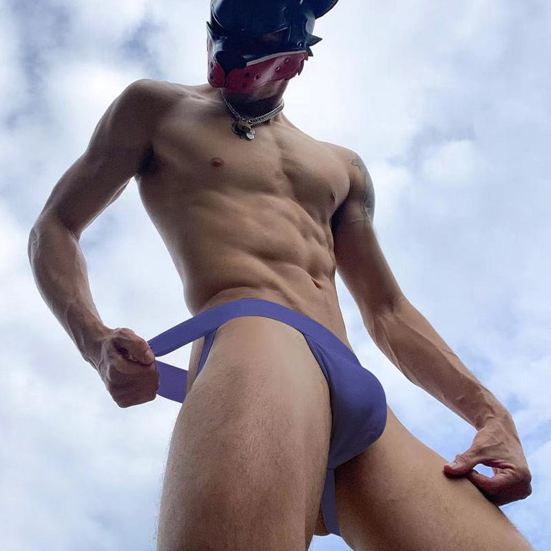 Low-angle model wearing a purple jockstrap and black leather mask outdoors