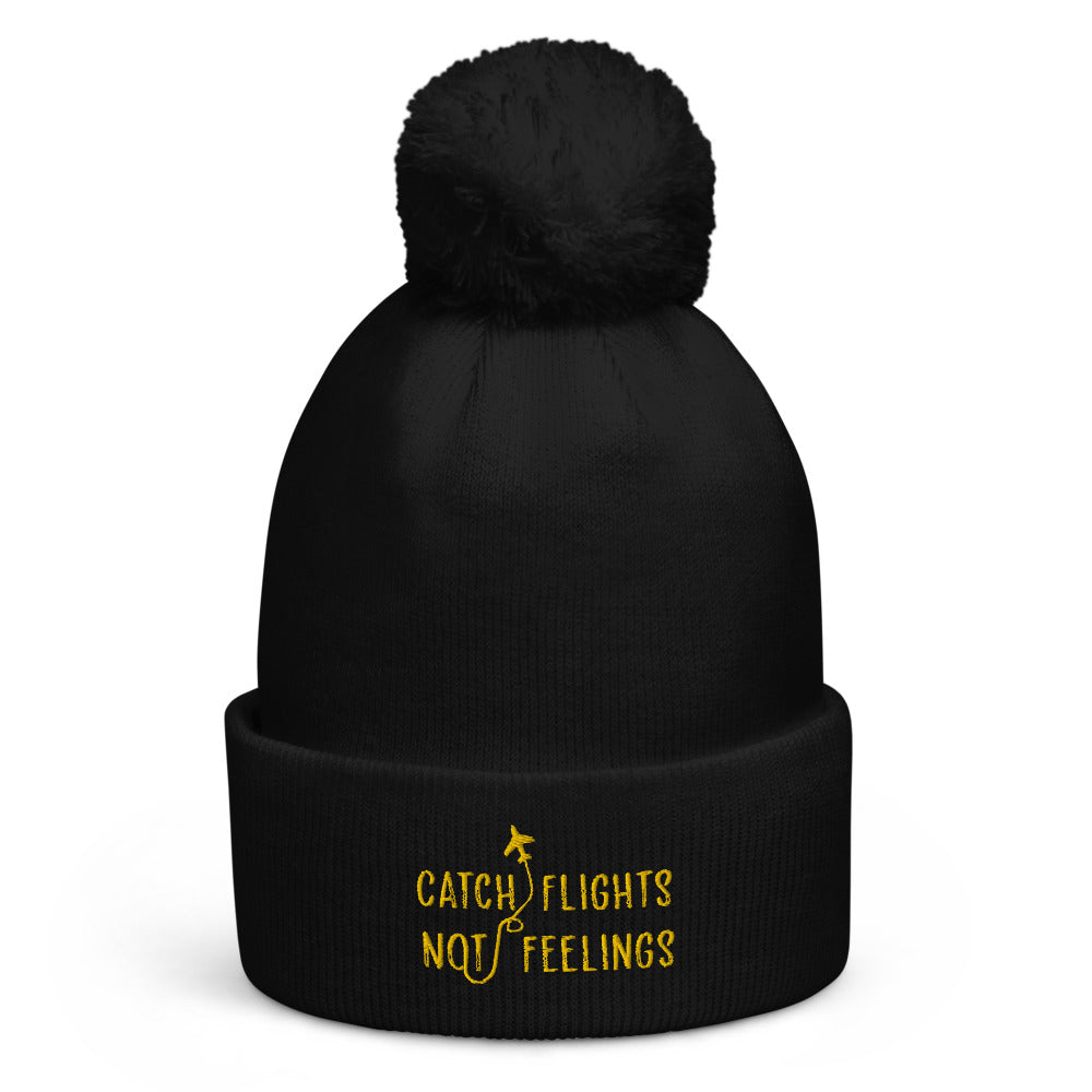 Black pom-pom beanie with yellow “CATCH FLIGHTS NOT FEELINGS” embroidery