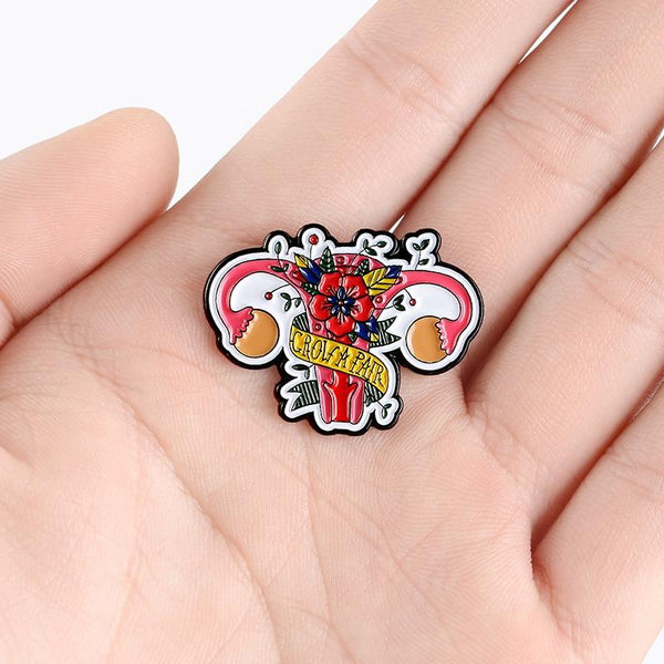 Hand holding a colorful enamel pin shaped like a flower-covered uterus with “GROW A PAIR” text