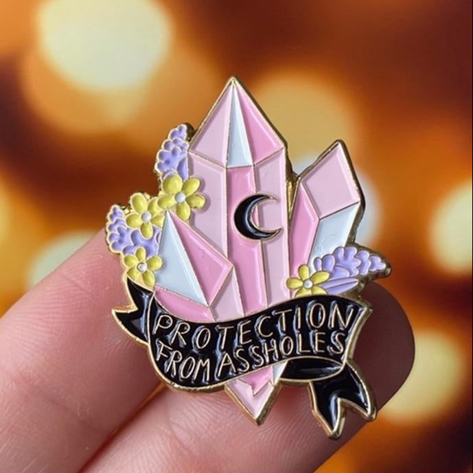 Pink crystal enamel pin reading “Protection From Assholes,” with floral accents