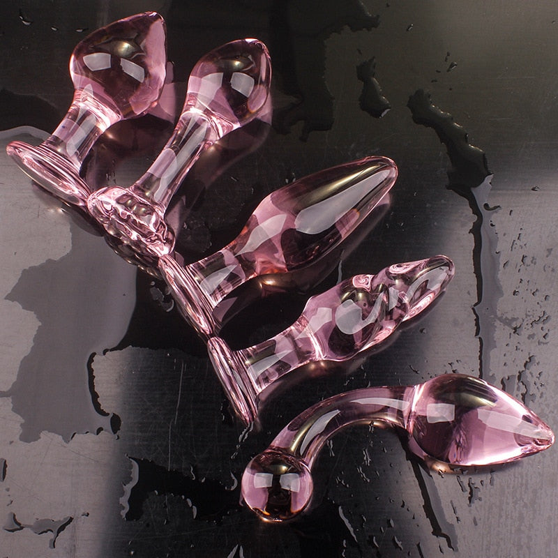 Five translucent pink glass butt plugs arranged on a wet dark surface