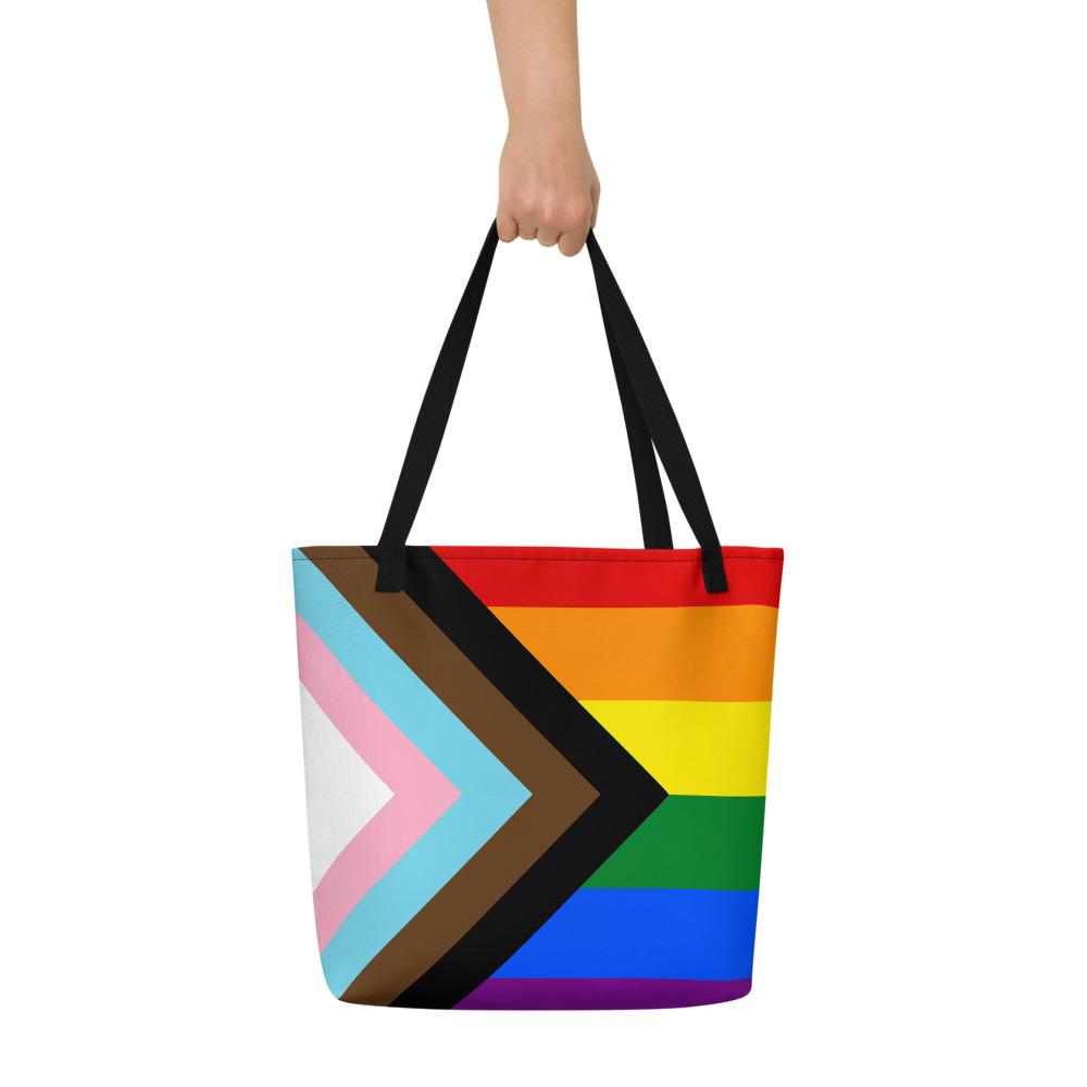 Black-handled tote bag held up, displaying a colorful Progress Pride flag design