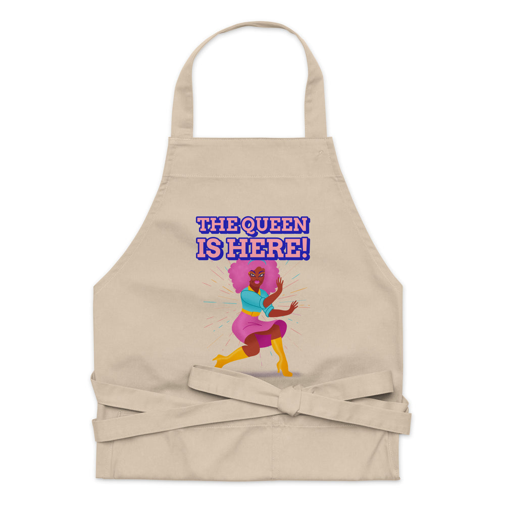 Front view of a beige apron reading “THE QUEEN IS HERE!” with a colorful illustrated queen