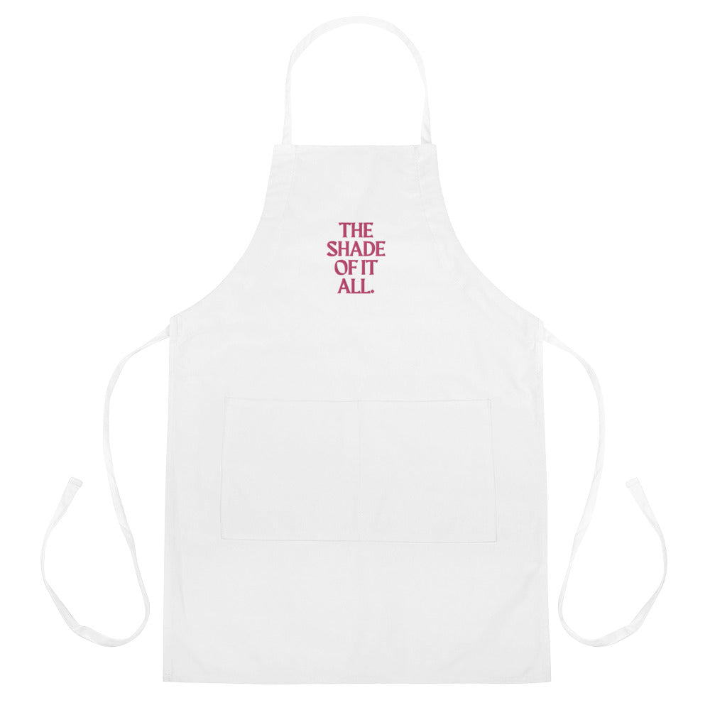 White apron with neck and waist ties, front pocket, and embroidered “THE SHADE OF IT ALL.”