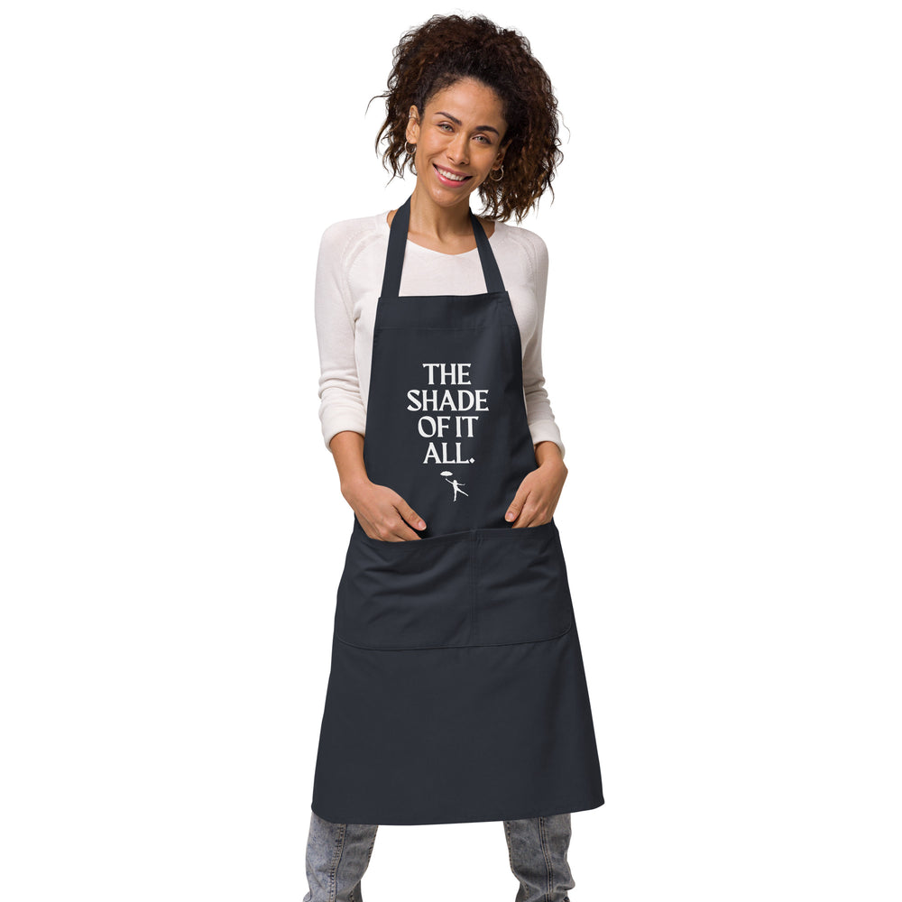 Front view of a black apron on a smiling model, reading “THE SHADE OF IT ALL.”