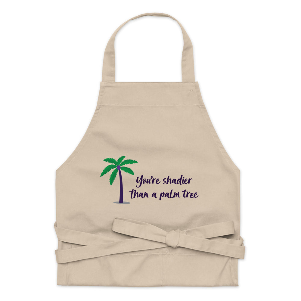 Front view of a beige apron with a palm tree and “You’re shadier than a palm tree”