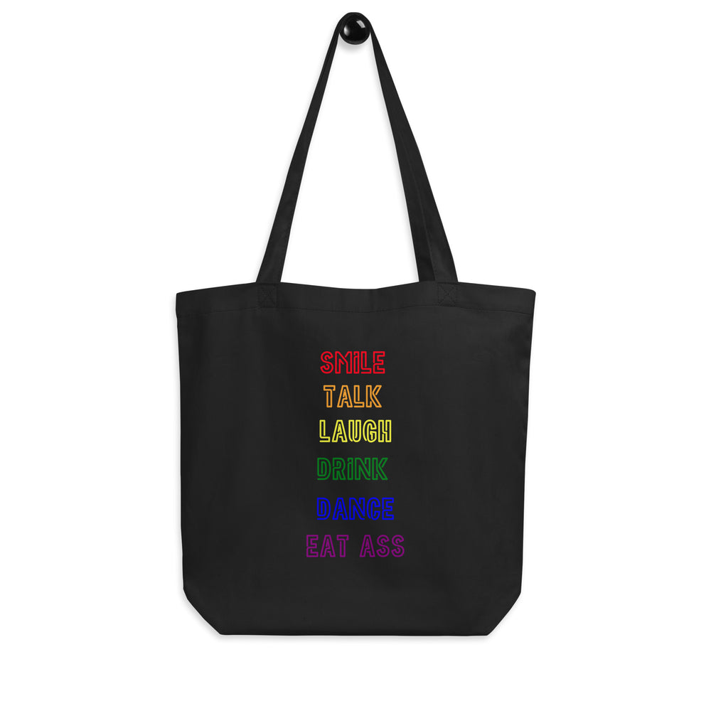 Front-facing black tote bag with rainbow text reading “SMILE TALK LAUGH DRINK DANCE EAT ASS”