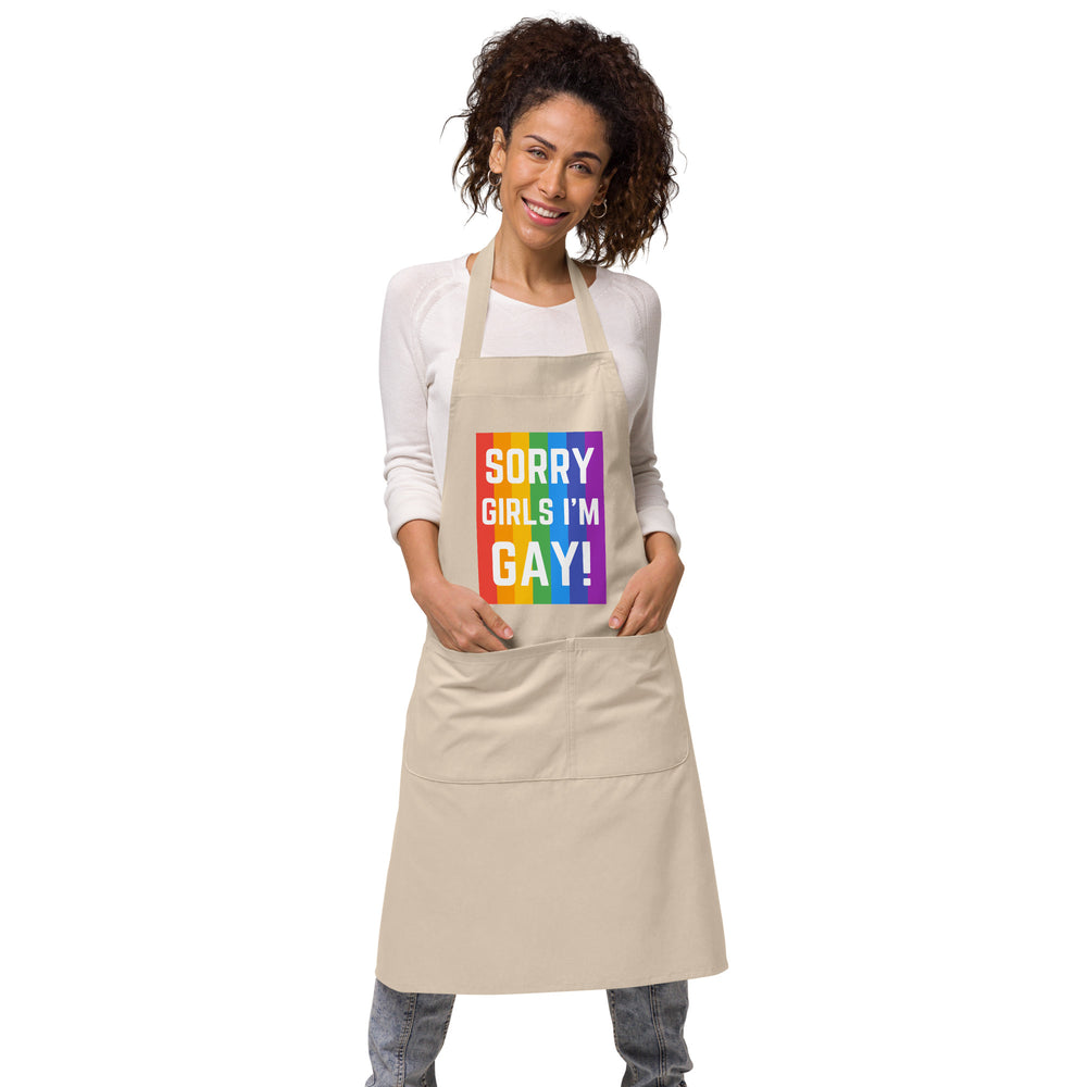 Front view of a beige apron on a smiling model, reading “SORRY GIRLS I’M GAY!” over rainbow stripes