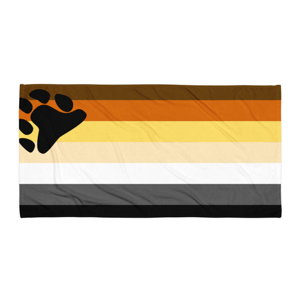 Beach towel with bold bear pride stripes and a black bear paw at the corner, shown flat on wood beside leather and stone accents.