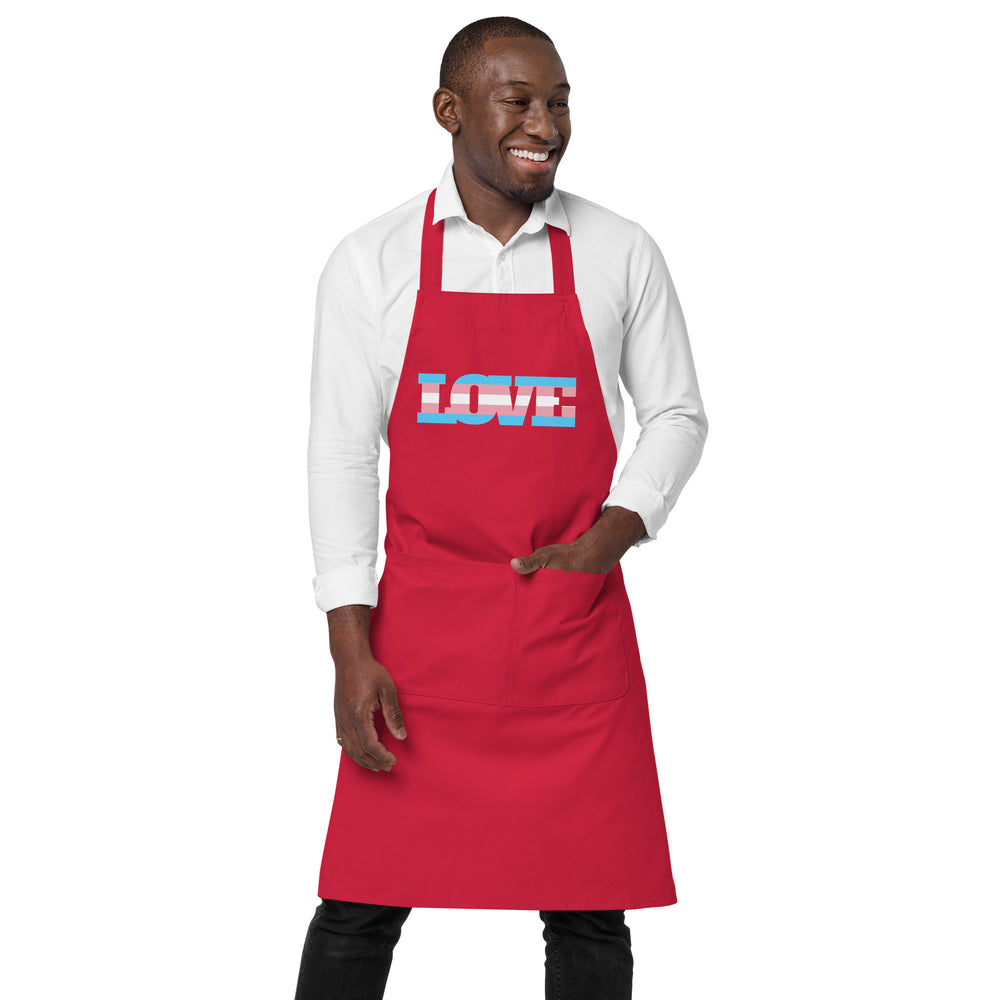 Front view of a smiling person wearing a red apron with a transgender pride stripe and LOVE text
