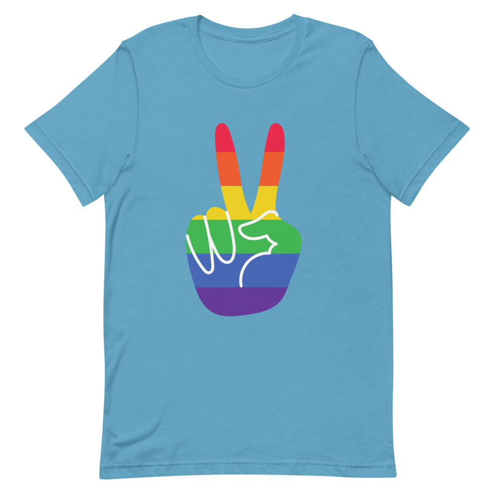 Gay Pride T-shirt featuring a rainbow peace sign, worn by a smiling person with shoulder-length dark hair in a cozy living room.