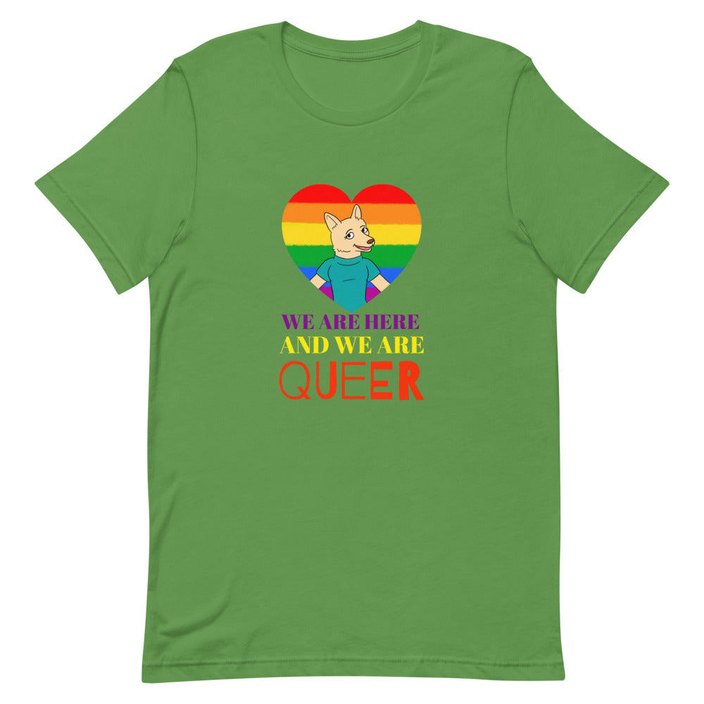 We Are Here And We Are Queer" T-shirt featuring a cartoon cat and rainbow-striped heart, worn by a smiling queer person indoors.
