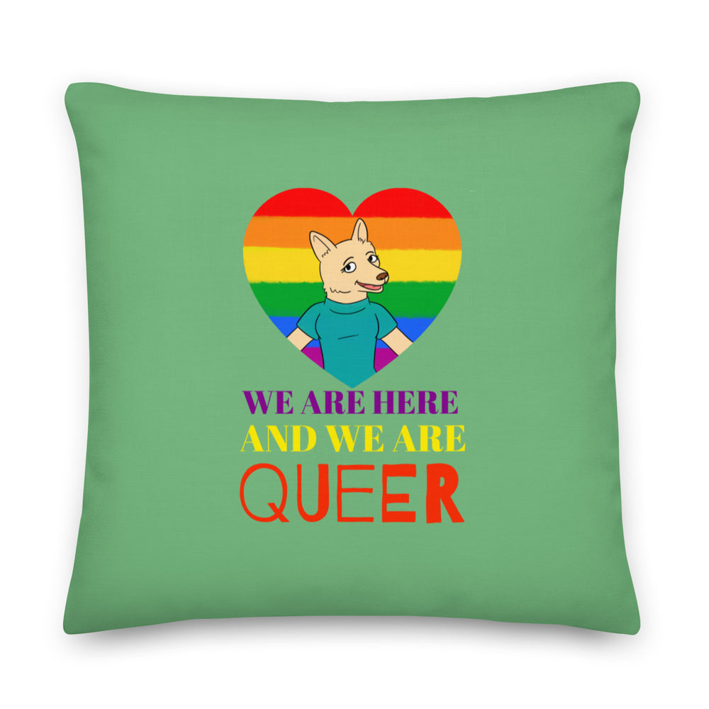 Square green pillow with a rainbow heart, cartoon canine, and “WE ARE HERE AND WE ARE QUEER” text