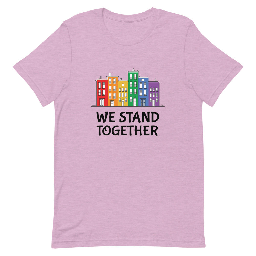 Front view of a heather lilac T-shirt reading “WE STAND TOGETHER” below colorful buildings
