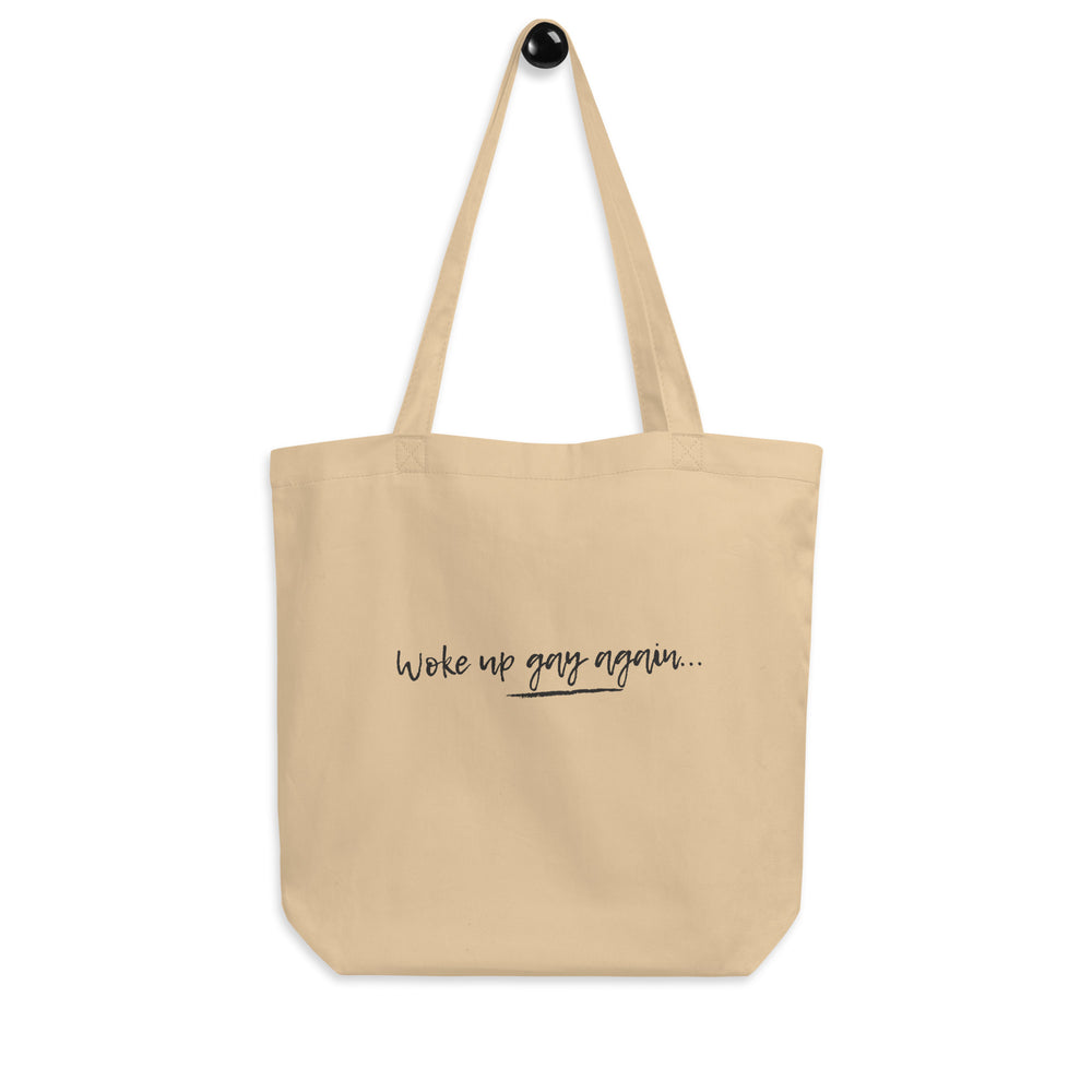 Front-view beige tote bag reading “Woke up gay again...” in black script