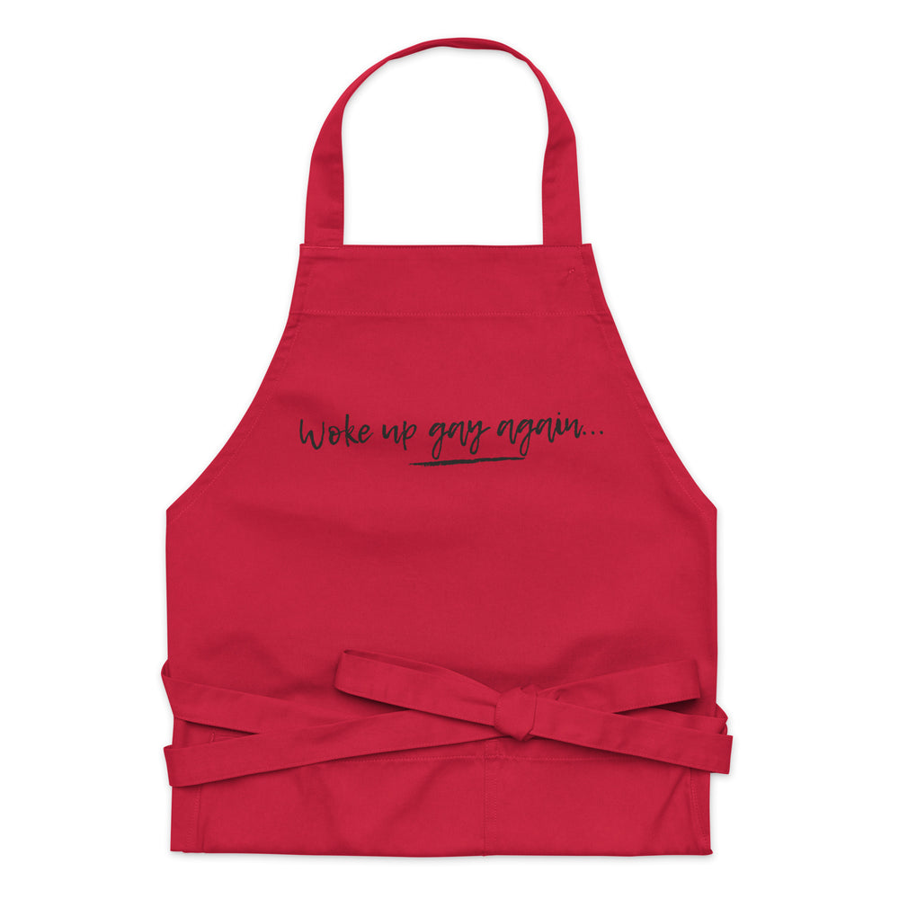 Front-facing red apron with black script reading “Woke up gay again...” and waist ties