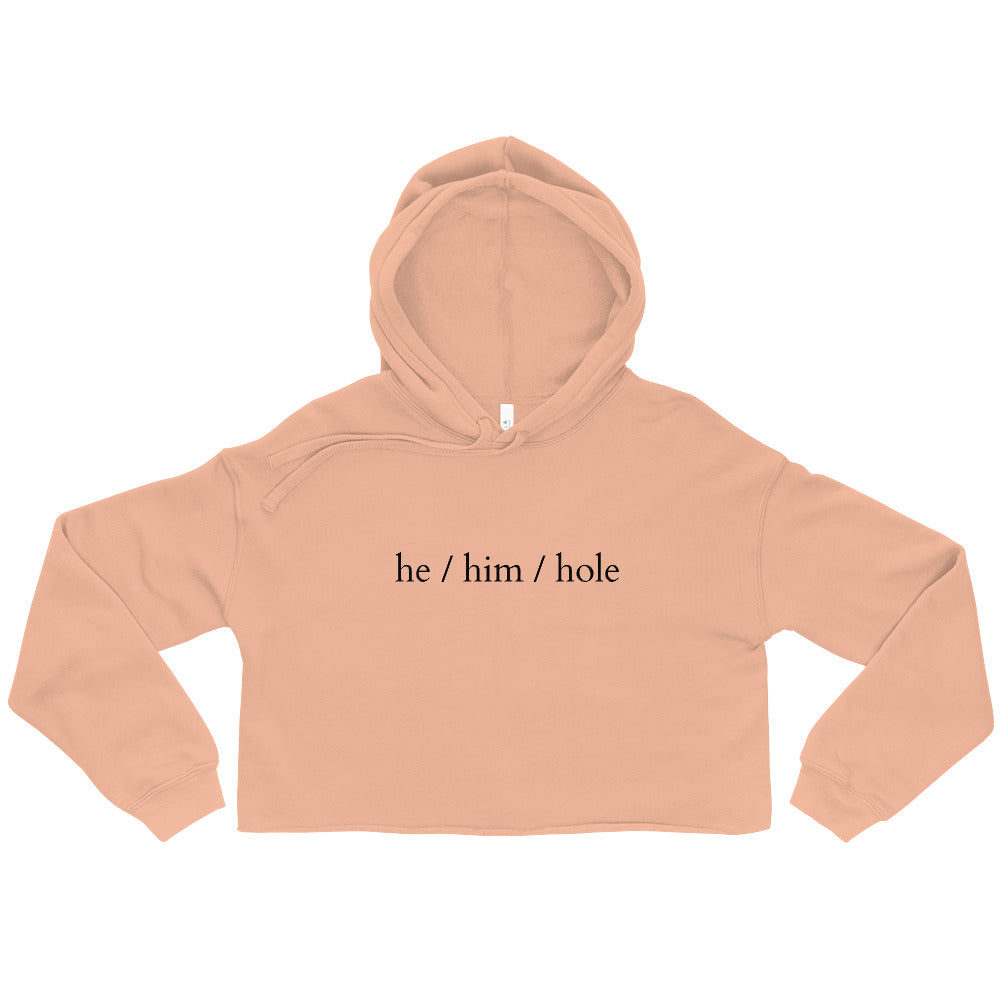 Peach cropped hoodie laid flat, printed with “he / him / hole” in black