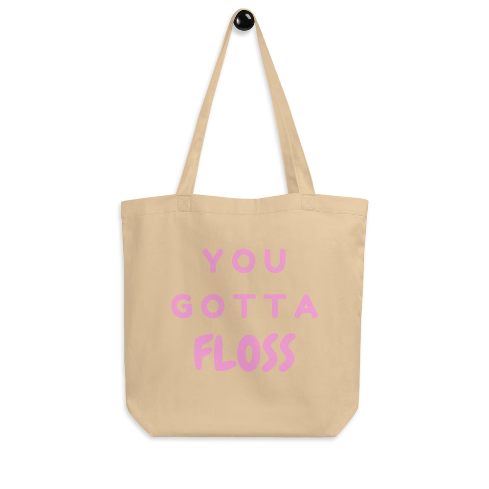 Front view of a beige tote bag with pink lettering reading “YOU GOTTA FLOSS.”